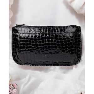 May include: A rectangular black clutch bag featuring a glossy, patent finish with a textured crocodile skin pattern. The bag has a slim profile, rounded corners, and a silver-toned metal zipper closure running along the top edge of the accessory.