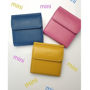 May include: Three small, square wallets in blue, pink, and yellow. Each wallet has a flap closure and a textured surface. The word "mini" is written in purple and green around the wallets.