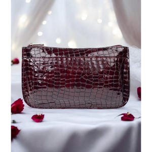 May include: A rectangular clutch bag in a deep burgundy color with a glossy, patent finish. The exterior features a textured, embossed crocodile skin pattern and a silver-toned metal zipper closure along the top edge of the accessory.