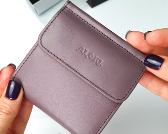 May include: A square, mauve-colored leather coin purse featuring a flap closure with the embossed text ALORI. The wallet has visible tonal stitching along the edges of the flap and the main body, presenting a smooth, matte finish throughout the exterior.