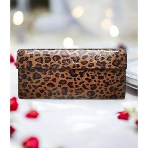 Puede incluir: Una cartera rectangular con estampado de leopardo marrón y negro. La cartera tiene una solapa y un acabado brillante. El fondo está borroso, con iluminación suave y pétalos de rosas rojas.