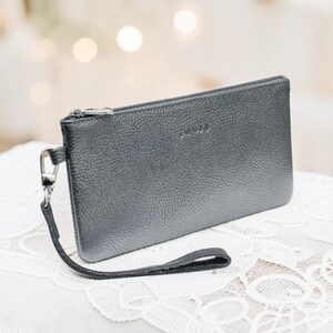 Könnte beinhalten: Eine rechteckige, dunkelgraue Handgelenktasche mit genarbter Textur, ausgestattet mit einem silberfarbenen Reißverschluss an der Oberseite und einer passenden abnehmbaren Handschlaufe, die mit einem silbernen Karabinerhaken befestigt ist.