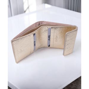 Può includere: Portafoglio dorato aperto con finitura strutturata. Il portafoglio è aperto, rivelando fessure per carte e un interno rosa chiaro. Il nome del marchio "ALORI" è visibile sugli slot per carte. Il portafoglio è su una superficie bianca.