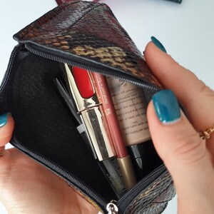 A small multi coloured zippered pouch with a snake print. The pouch is open and contains three makeup items: a red lipstick, a nude lipstick, and a black eyeliner.