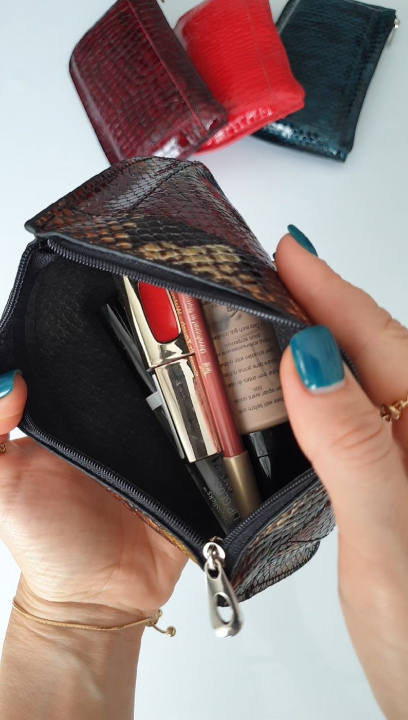 A small multi coloured zippered pouch with a snake print. The pouch is open and contains three makeup items: a red lipstick, a nude lipstick, and a black eyeliner.