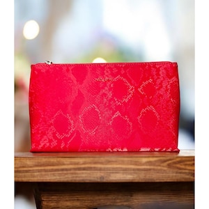 May include: A rectangular red cosmetic bag with a snakeskin pattern. The bag has a silver zipper and a small logo at the top. The bag is sitting on a wooden surface.