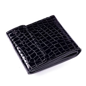 A black, glossy wallet with a crocodile skin texture. The wallet is folded and has a snap closure. The surface reflects light, highlighting the texture and shape of the wallet. The wallet to made of leather.