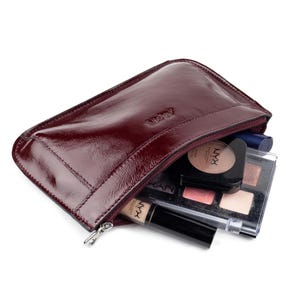 May include: A burgundy leather cosmetic bag with a silver zipper, partially open to reveal makeup products. The bag contains a compact, eyeshadow palette, and a tube of concealer. The bag has the word "Misty" embossed on the top.