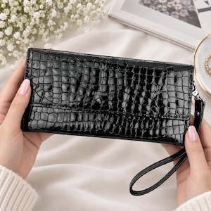 Op de afbeelding: Een rechthoekige zwarte clutch met een glanzende afwerking en een gestructureerd krokodillenpatroon. De portemonnee is voorzien van een bijpassende zwarte polsband die aan de zijkant is bevestigd met een zilverkleurige metalen sluiting en een horizontale klepsluiting.