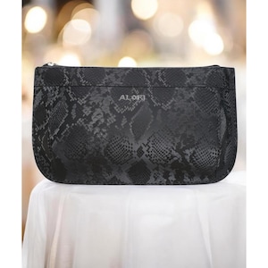 May include: Black clutch with a snakeskin pattern. The rectangular bag has a zipper closure and the word "ALORI" printed on the front. The bag is sitting on a white surface.