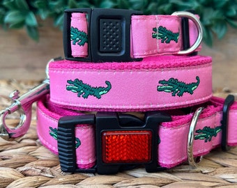 May include: Pink dog collar and leash set with a green alligator print. The collar has a black buckle and a reflective red safety tag. The leash has a silver clip.