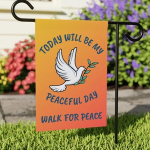 May include: A garden flag with a dove carrying an olive branch, set against an orange and yellow gradient background. The flag reads "Today Will Be My Peaceful Day Walk For Peace" in blue text. The flag is displayed on a black metal stand.