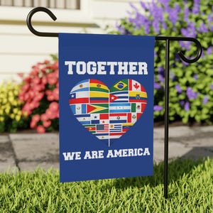 May include: A blue garden flag with the words "TOGETHER WE ARE AMERICA" in white. The flag features a heart shape filled with various national flags. The flag is displayed on a black metal stand in a grassy yard, with flowers in the background.
