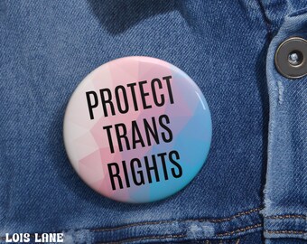May include: A pink, blue, and white button with the text "PROTECT TRANS RIGHTS" in black.