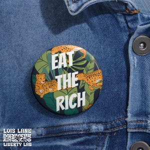 May include: A round button with a black background and a jungle print featuring green leaves and orange leopards. The button says "Eat the Rich" in white text.
