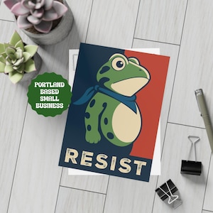 May include: A greeting card featuring a cartoon frog with a blue scarf, set against a red and navy blue background. The word "RESIST" is printed in a vintage font. The card is from a Portland-based small business.