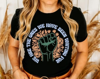 May include: Black t-shirt with a graphic print of a raised fist surrounded by flowers. The text "We are the ones we have been waiting for" is printed in a circle around the fist.