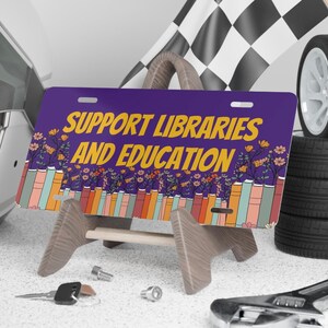 May include: Purple and white license plate with the text "Support Libraries and Education" in yellow. The plate is decorated with colorful flowers and books.