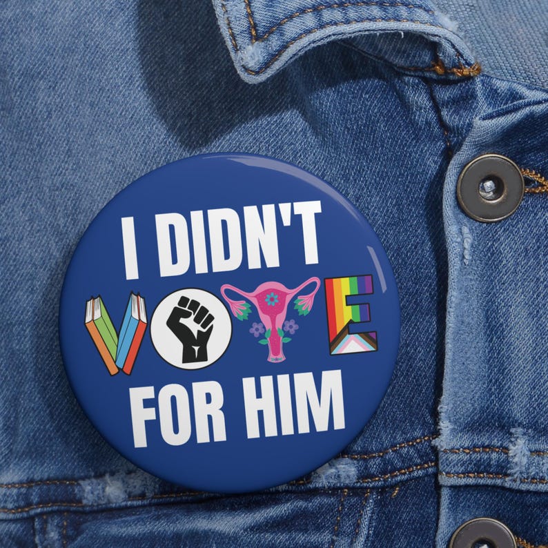 Blue button with white writing I DIDN'T VOTE FOR HIM with word VOTE made up of illustrations denoting anti book banning, Black Lives Matter, womens reproductive rights supporter, and LGBTQIA plus ally.