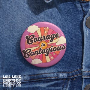May include: A round, pink button with the words "Courage is Contagious" in a retro font. The button features a sunburst design with orange, peach, and cream colors, along with white clouds and small hearts. The button is set against a blue denim background.