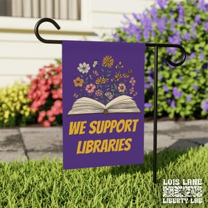May include: Purple garden flag with an open book and flowers. The text "We Support Libraries" is printed in yellow on the flag.