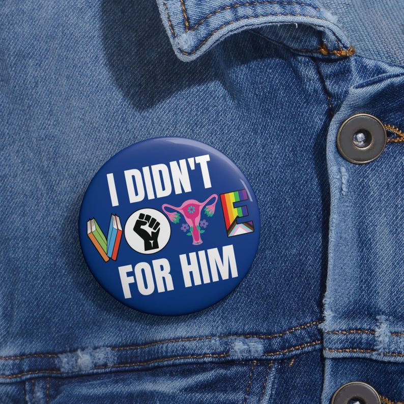 Blue button with white writing I DIDN'T VOTE FOR HIM with word VOTE made up of illustrations denoting anti book banning, Black Lives Matter, womens reproductive rights supporter, and LGBTQIA plus ally.