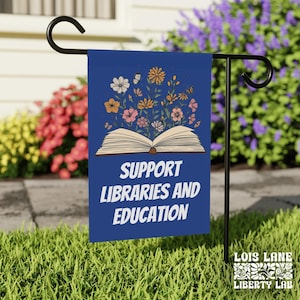 May include: A blue garden flag with the text "SUPPORT LIBRARIES AND EDUCATION" and an illustration of an open book with flowers and butterflies. The flag is displayed on a black metal stand in a garden.