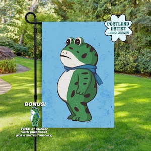 May include: A rectangular garden flag featuring a light blue background with a green frog illustration wearing a blue scarf. The flag includes a white flower-shaped badge with the text PORTLAND ARTIST HAND DRAWN and a small frog sticker graphic.
