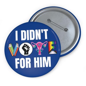 Blue button with white writing I DIDN'T VOTE FOR HIM with word VOTE made up of illustrations denoting anti book banning, Black Lives Matter, womens reproductive rights supporter, and LGBTQIA plus ally.