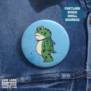 May include: A round button with a cartoon frog design. The frog is green with dark green spots and a blue scarf, set against a light blue background. Text on the button reads "Portland Based Small Business". The button is on a denim jacket.