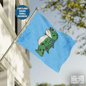 May include: A light blue flag featuring a cartoon frog with green spots, a white belly, and a blue scarf. The flag has a distressed look and the text "Portland Based Small Business". The flag is mounted on a silver pole.