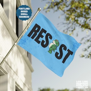 May include: A light blue flag with the word "RESIST" in large black letters and a cartoon frog. The flag also features the text "Portland Based Small Business". The flag is mounted on a silver pole.