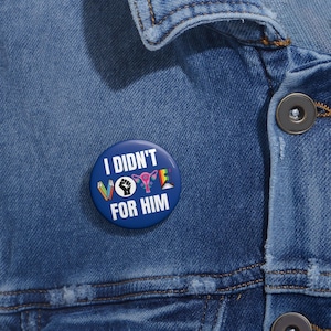 Blue button with white writing I DIDN'T VOTE FOR HIM with word VOTE made up of illustrations denoting anti book banning, Black Lives Matter, womens reproductive rights supporter, and LGBTQIA plus ally.