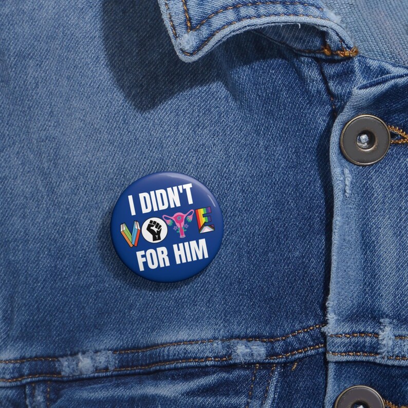 Blue button with white writing I DIDN'T VOTE FOR HIM with word VOTE made up of illustrations denoting anti book banning, Black Lives Matter, womens reproductive rights supporter, and LGBTQIA plus ally.