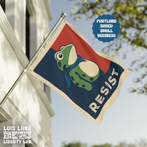 May include: A flag with a cartoon frog wearing a blue scarf, set against a red and navy blue background. The word "RESIST" is printed in white. The flag also includes the text "Portland Based Small Business" and the logo for "Lois Lane Liberty Lab".