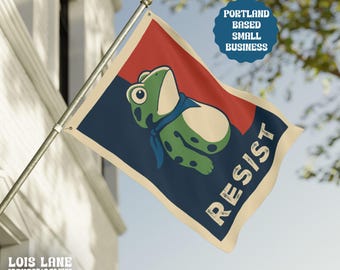 May include: A flag with a cartoon frog wearing a blue scarf, set against a red and navy blue background. The word "RESIST" is printed in white. The flag also includes the text "Portland Based Small Business" and the logo for "Lois Lane Liberty Lab".