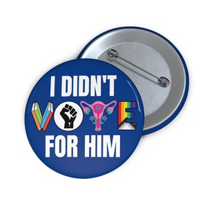 Blue button with white writing I DIDN'T VOTE FOR HIM with word VOTE made up of illustrations denoting anti book banning, Black Lives Matter, womens reproductive rights supporter, and LGBTQIA plus ally.