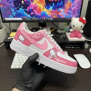 May include: A white and pink Nike Air Force 1 sneaker featuring Hello Kitty graphics. The shoe has a pink swoosh, pink accents, and the name "Ayanna" printed on the side. The shoe is held by a gloved hand.