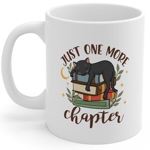 White ceramic coffee cup featuring a whimsical design of a magical stack of books with a black cat sleeping on top. The text 'Just One More Chapter' is displayed above and below the image. Available in 11oz and 15oz sizes, with a C-shaped easy-grip handle. The mug has a glossy finish and is lead and BPA-free.
