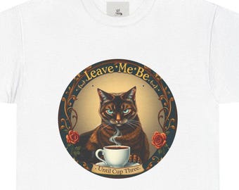 Op de afbeelding: Een wit T-shirt met een vintage-achtige illustratie van een kat die een kopje koffie vasthoudt. De illustratie is omgeven door een ronde lijst met de tekst "Leave Me Be. Until Cup Three."