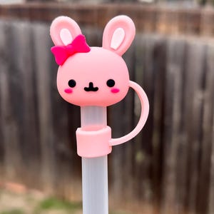 May include: Pink bunny-shaped pen topper with a pink bow. The topper has a white face with black eyes and a black nose. The topper is attached to a white pen.