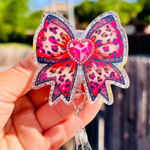 May include: A pink and black leopard print bow with a large pink rhinestone in the center. The bow is attached to a clear plastic retractable badge holder.