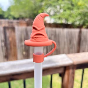 May include: An orange-colored, wizard hat-shaped straw topper. The topper is made of a soft material and features a detailed design. The straw is white and the topper has a small handle. The background is a blurred outdoor scene.