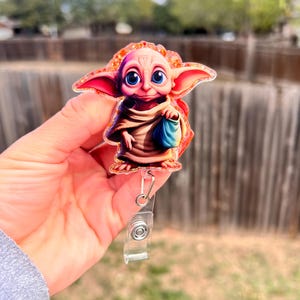 May include: A badge reel featuring a cartoon character with large blue eyes, large ears, and a tan robe. The badge reel has a clear plastic retractable cord and a metal clip. The character is holding a small blue bag.