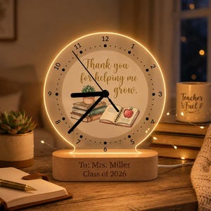 Könnte beinhalten: Eine kreisförmige Acryl-Tischuhr mit warmem LED-Lichtrand, die den Text Thank you for helping me grow, einen Bücherstapel, eine Sukkulente und ein offenes Buch mit einem Apfel zeigt. Der Holzsockel ist mit To: Mrs. Miller Class of 2026 graviert.