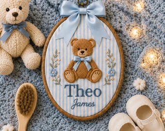 May include: An oval wooden embroidery hoop featuring a light blue and white striped fabric background with a central brown teddy bear appliqué, a light blue satin bow, and the embroidered name Theo James in blue thread, flanked by floral sprigs.
