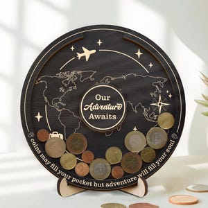 May include: A circular black wooden coin bank with a world map engraving, a central circle reading "Our Adventure Awaits," and a bottom border text "coins may fill your pocket but adventure will fill your soul." Includes an aeroplane and compass icon.