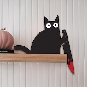 Peut inclure: Un présentoir en forme de chat noir avec des yeux blancs et un couteau à manche rouge. Une citrouille rose et un livre intitulé "MAGNOLIA TABLE" sont posés sur une étagère en bois contre un mur à panneaux blancs. Décoration sur le thème d'Halloween.