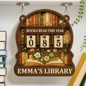 Puede incluir: Un letrero de madera con el texto "BOOKS READ THIS YEAR" y los números "085". El letrero tiene un diseño de estantería con un libro abierto y detalles florales. El texto "EMMA'S LIBRARY" está en la parte inferior.
