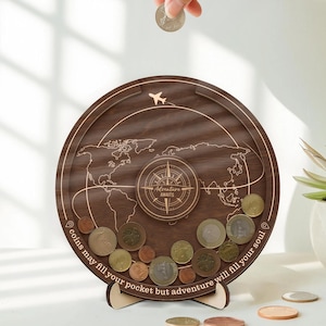 May include: A wooden travel-themed coin bank with a world map design, a compass, and the text "Adventure Awaits." The bank is filled with various coins, and a hand is dropping a coin into the slot. The bank is supported by a wooden stand.